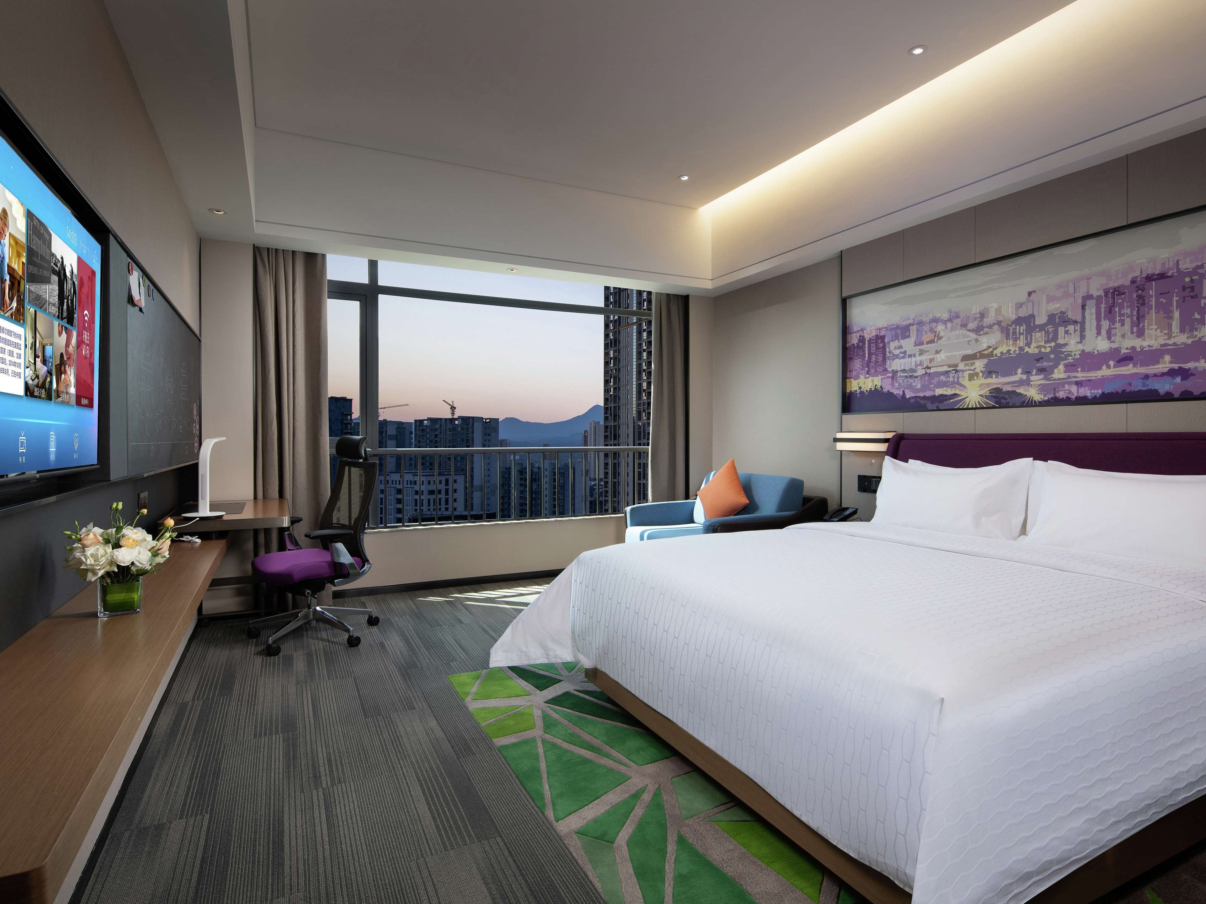 Une chambre d’hôtel moderne dotée d’un grand lit, d’un bureau avec un ordinateur, d’une chaise, d’une télévision et d’une grande fenêtre donnant sur la ville.