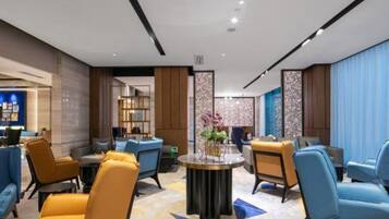 A modern hotel lobby with a central round table, seating areas with blue and yellow chairs, and decorative elements.