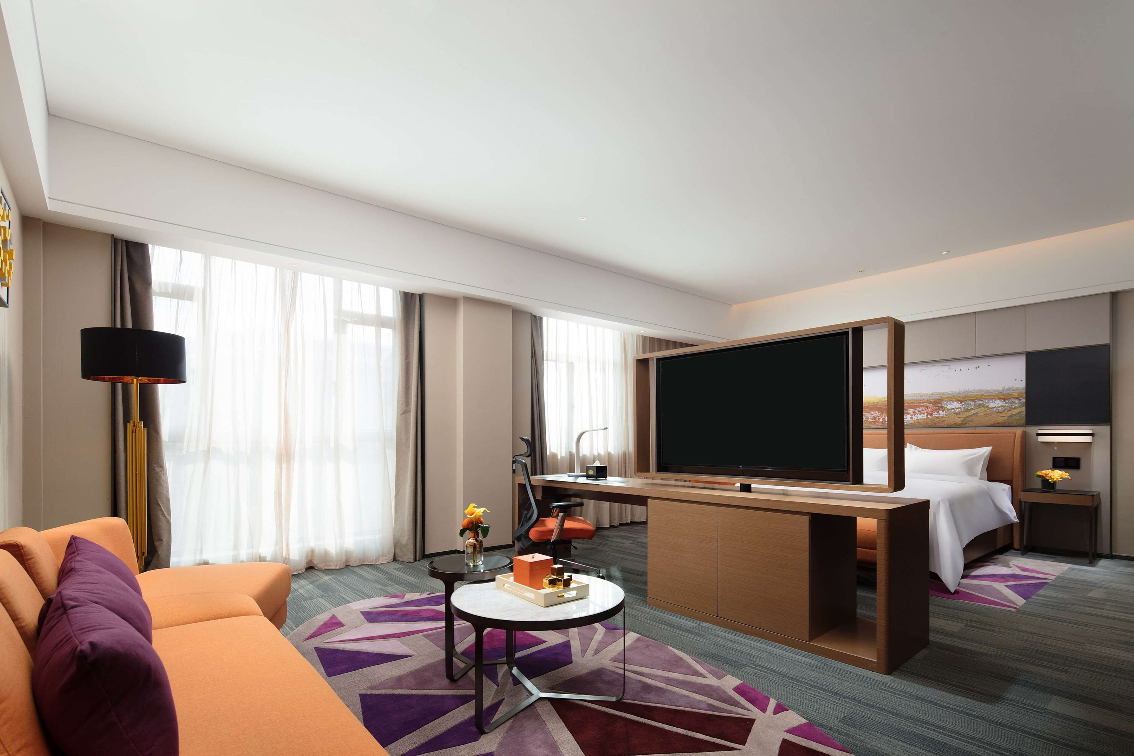Quarto de hotel com cama, escrivaninha, televisão, sofá e janela com cortinas.