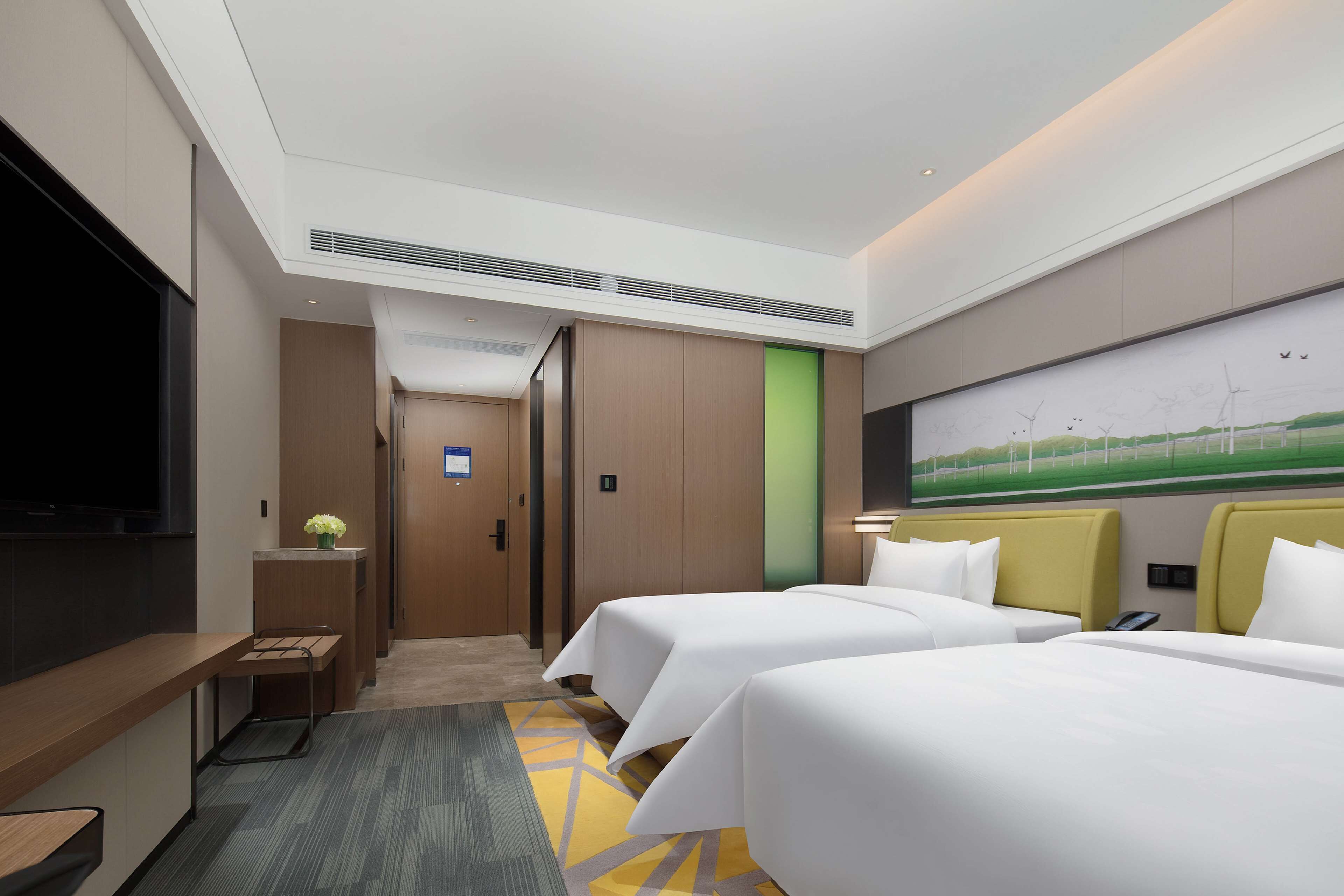 A hotel room with two beds, a desk, a TV, and a painting of wind turbines.