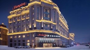 Um hotel grande, de vários andares, com uma placa 'Hampton' bem visível, apresentando um estilo arquitetônico clássico com janelas simétricas e uma entrada central.