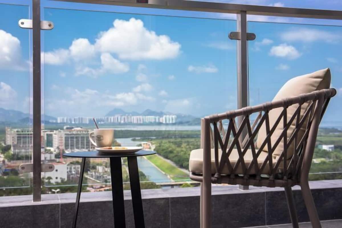 A balcony with a view of a river and mountains, a table with a cup and saucer, and a wicker chair.