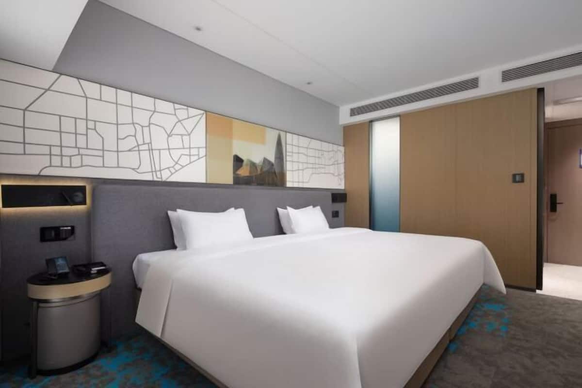 A hotel room with a large bed, a bedside table, a trash bin, and a wall with a cityscape design.