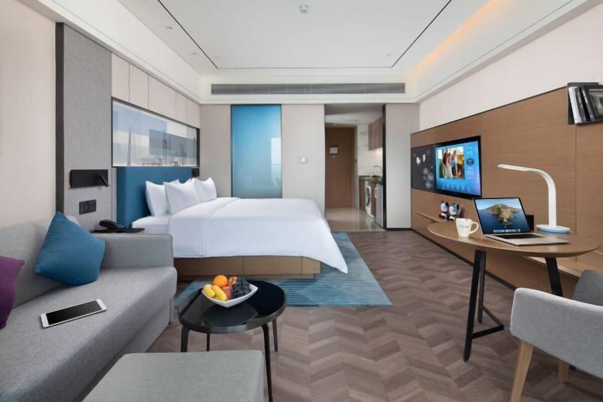 A modern hotel room with a bed, a desk with a laptop, a sofa, and a small table with fruits.