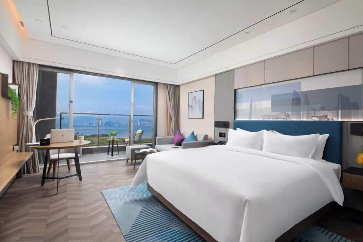 Une chambre d’hôtel moderne avec un grand lit, un bureau avec un ordinateur portable, un balcon avec vue et un tableau représentant un paysage urbain accroché au mur.