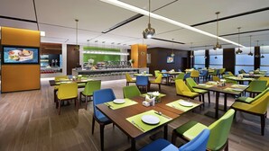 A modern restaurant with multiple dining tables and chairs, a central bar area, and a television displaying menu items.