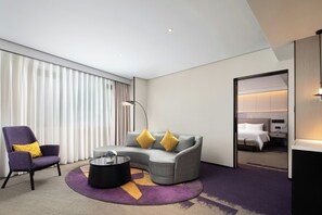 Una habitación de hotel moderna con un sofá gris curvo, un sillón morado, una mesa de centro negra y una cama con ropa de cama blanca.