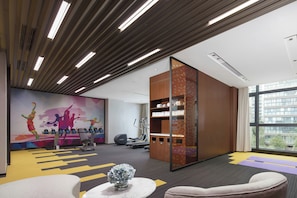 A modern gym with exercise equipment, a large mural, and a view of the city through large windows. - Hampton BY Hilton Qionghai Railway (QIONGHAI)