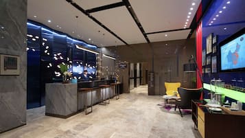 A modern hotel lobby with marble flooring, a reception desk, bar area, seating, and a television displaying a map.
