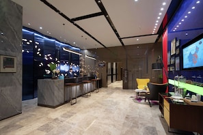 Un moderno vestíbulo de hotel con suelo de mármol, un mostrador de recepción, zona de bar, asientos y un televisor que muestra un mapa.