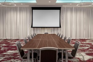 Uma sala de conferências com uma mesa grande, cadeiras, uma tela e cortinas.