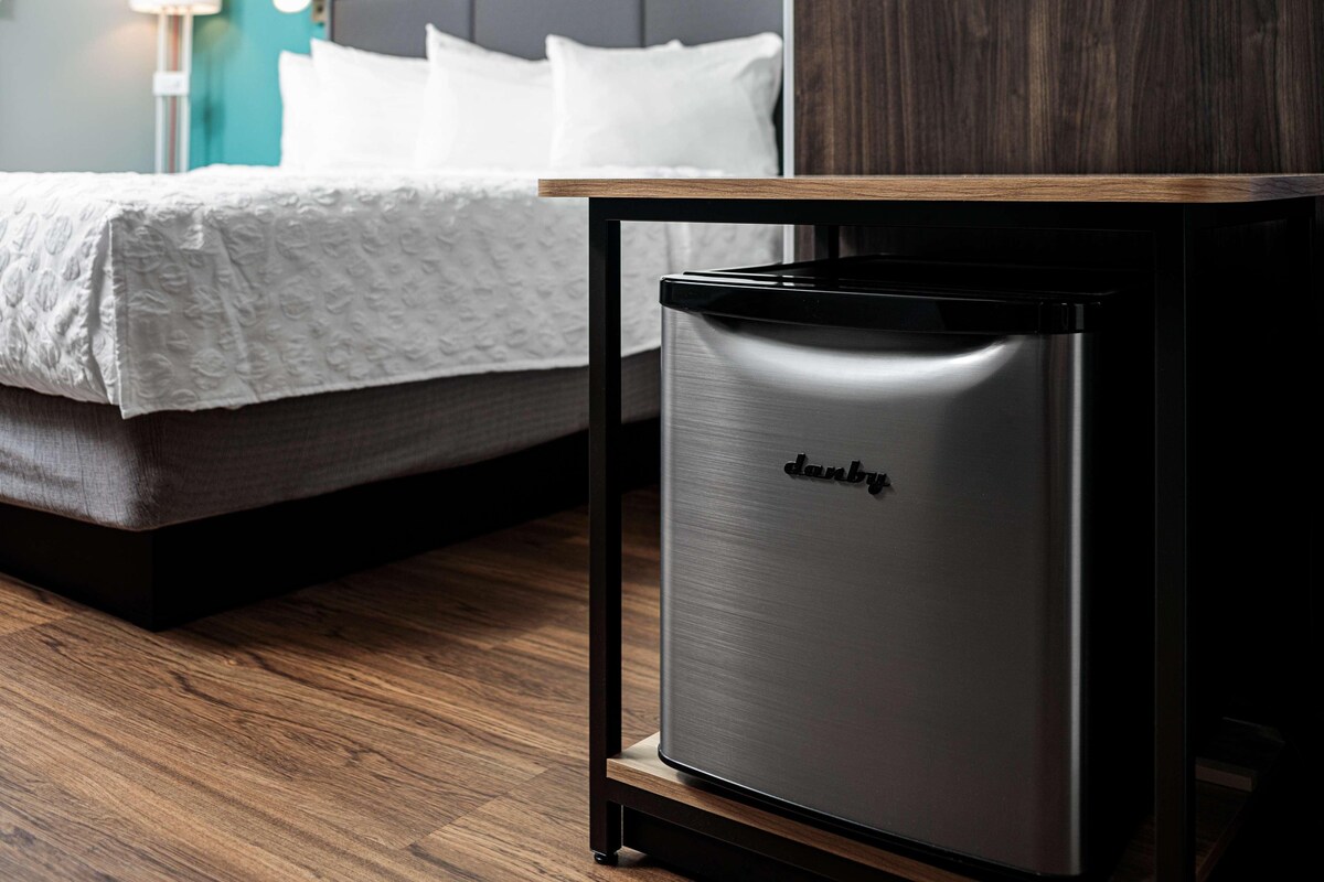 Ett kompakt kylskåp i rostfritt stål från Danby, placerat i ett modernt hotellrum med ett träbord, en säng med vita sängkläder och ett trägolv.