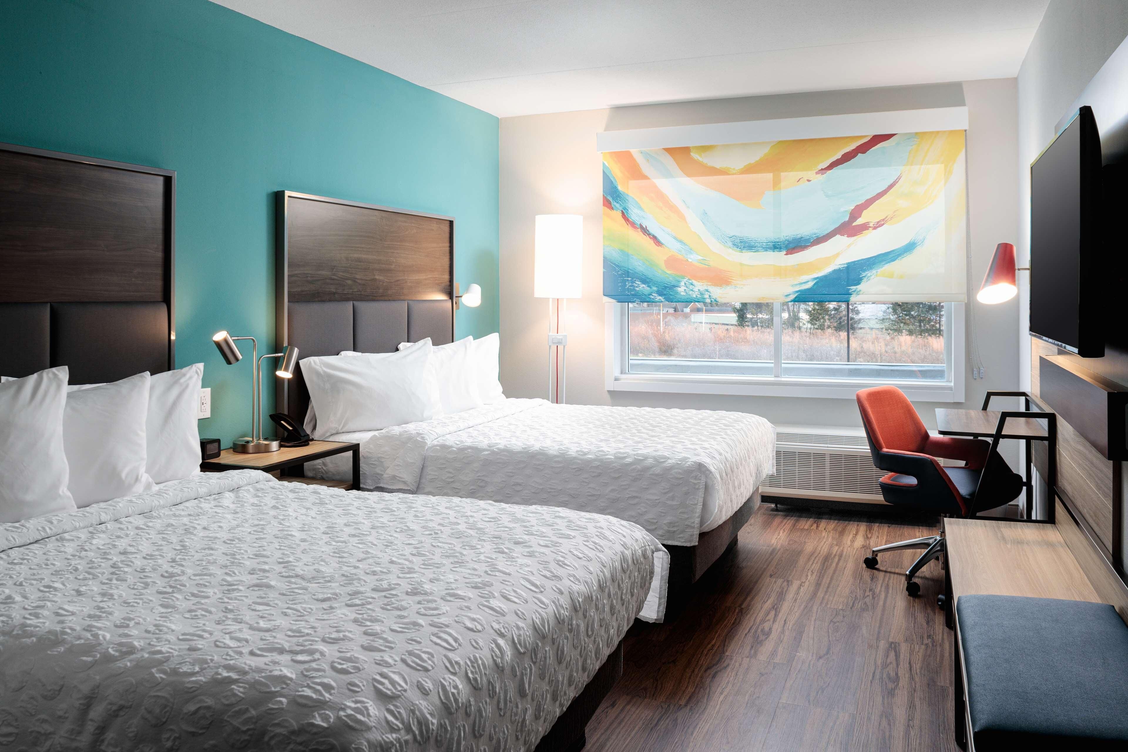 A hotel room with two beds, a desk, a chair, a TV, and a large window with a view.
