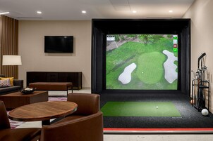 Quarto moderno com uma tela grande exibindo um campo de golfe, um carrinho de golfe e uma área de estar com mesa e cadeiras.