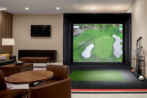Una habitación moderna con una pantalla grande que muestra un campo de golf, un carrito de golf y una zona de estar con una mesa y sillas.