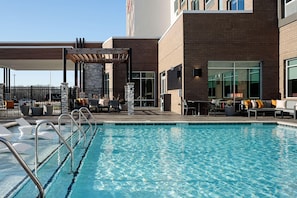 Un área de piscina moderna de hotel con una piscina cristalina, sillas de descanso y un espacio de estar cubierto.