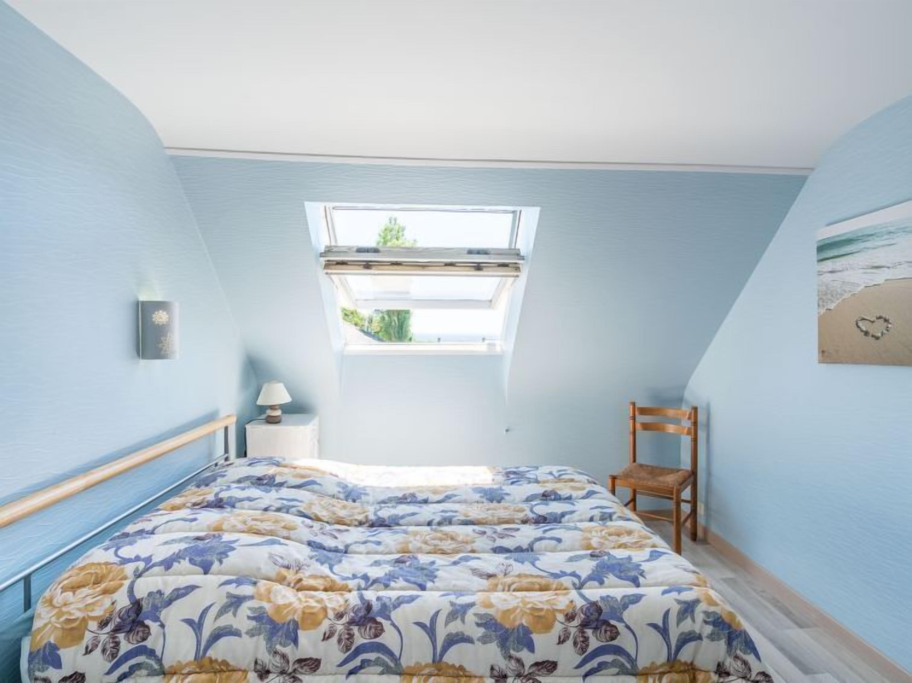 A bedroom with a skylight, a bed with floral bedding, a bedside table with a lamp, and a chair.