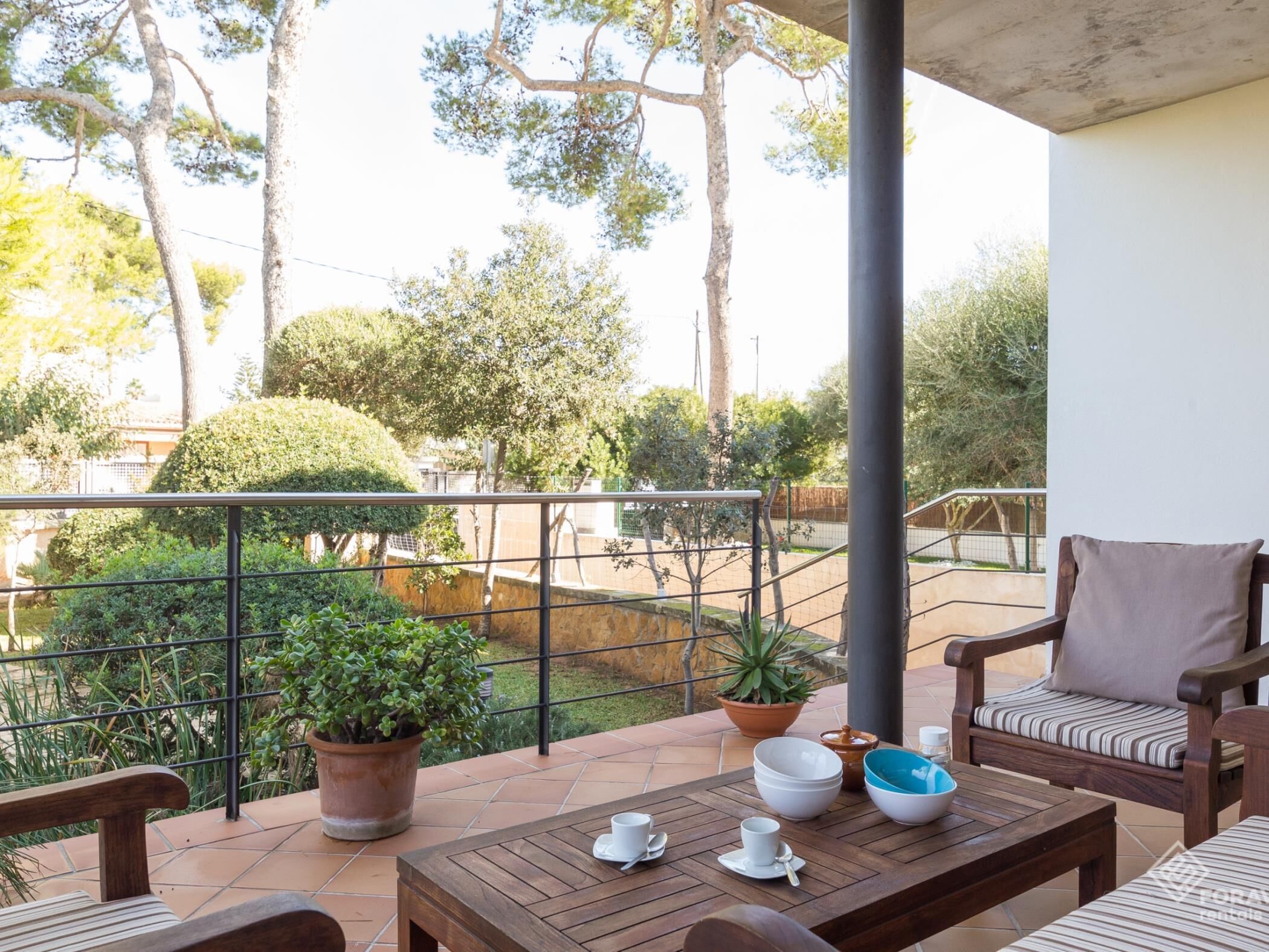 Ein Balkon mit Holztisch für zwei Personen, Blumentöpfen und einem Sessel mit Kissen.