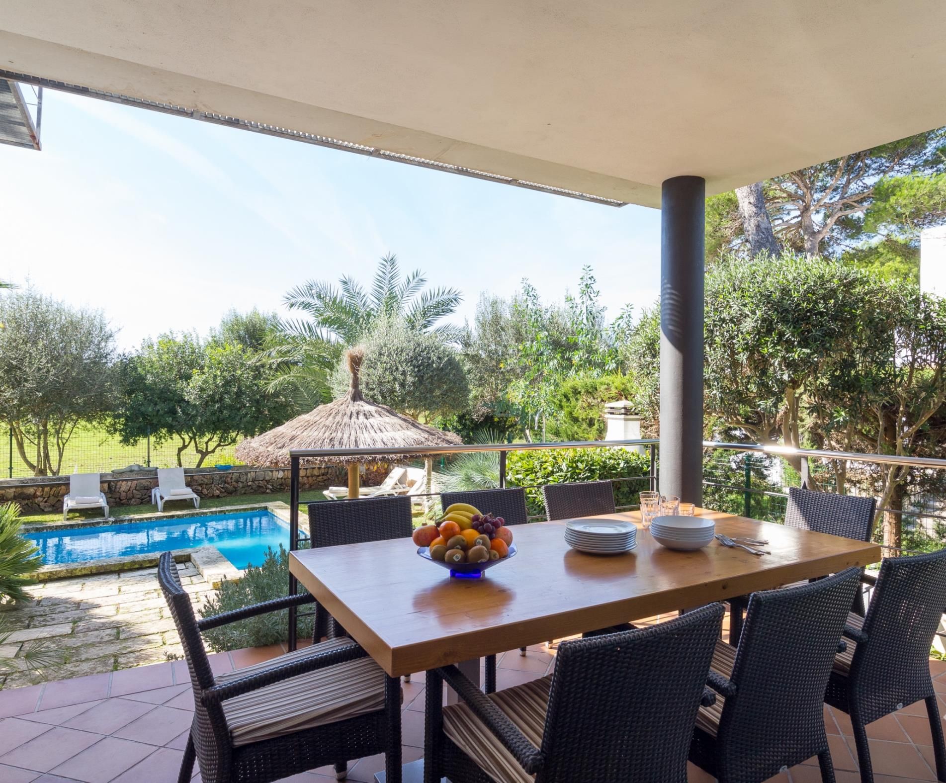 An outdoor dining area with a pool, wicker chairs, and a table with fruit and plates.