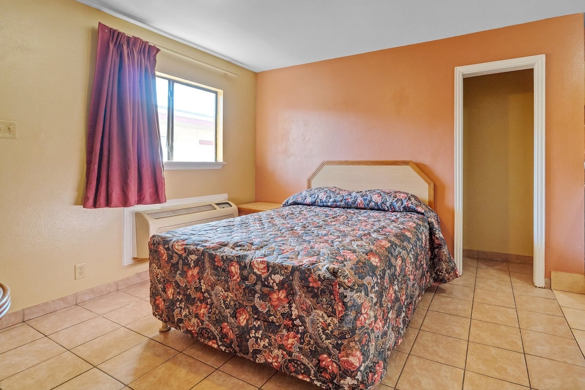 Un dormitorio con cama, ventana con cortinas, radiador y puerta a otra habitación.