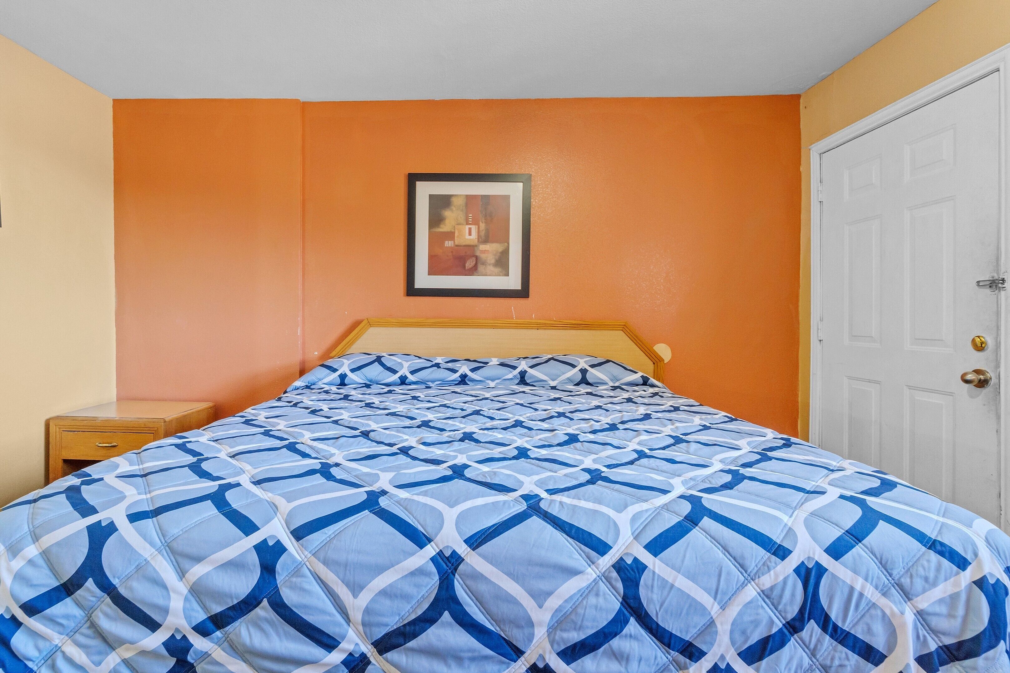 A bed with a geometric patterned bedspread, a wooden headboard, a nightstand with a drawer, and a framed picture on the wall.