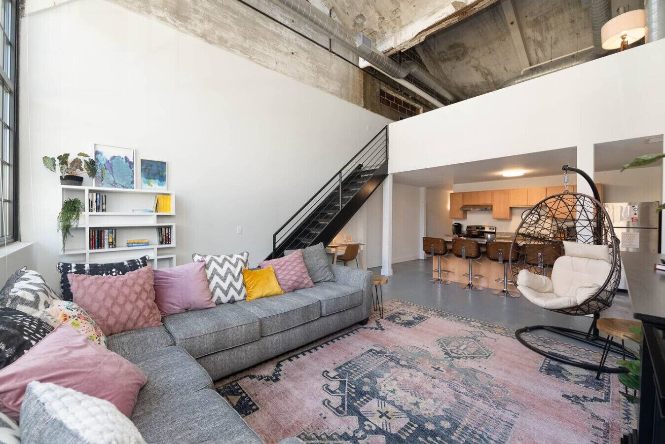 A modern living space with a grey sectional sofa, a hanging chair, a staircase, and a kitchen area.