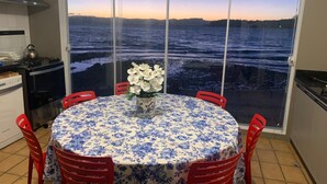 Una mesa redonda para comer con mantel floreado, rodeada de sillas rojas, frente a un amplio ventanal con vistas a un cuerpo de agua al atardecer.