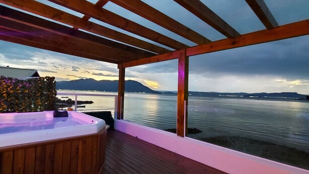 Una terraza de madera con jacuzzi con vistas a un lago, una pérgola de madera y vistas a montañas.