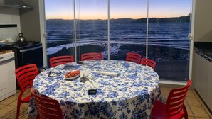 Un comedor con una mesa dispuesta para cuatro, vistas al mar a través de ventanas amplias y una zona de cocina con estufa y armarios.