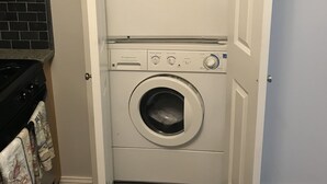 A compact, white laundry room with a front-loading washer and dryer, a small shelf above the dryer, and a tiled backsplash. - Oceanview Apt - Steps to the Beach! (Long Beach)