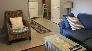 A living room with a wicker chair, a blue sofa, a coffee table with remote controls, a patterned rug, and a hallway leading to a bathroom. - Oceanview Apt - Steps to the Beach! (Long Beach)