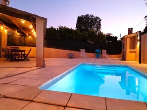 Una piscina rectangular con un borde de baldosas, rodeada de sillas de descanso y un porche cubierto con luces colgantes.