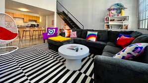 A modern living room with a black and white striped rug, a round coffee table, a black sofa with colorful cushions, a white hanging chair, and a staircase leading to an upper level. - 5109 Peters Cartridge Factory · ✪Kings Island Loft w/ Brewery, River, Bike trails✪ (Maineville)