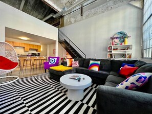 Een moderne woonkamer met een zwart-wit gestreept tapijt, een ronde salontafel, een zwarte bank met kleurrijke kussens, een witte hangstoel en een trap naar een hoger gelegen verdieping.