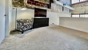 A room with a television on a dark cabinet, a patterned ottoman, and a floor lamp. - 5109 Peters Cartridge Factory · ✪Kings Island Loft w/ Brewery, River, Bike trails✪ (Maineville)
