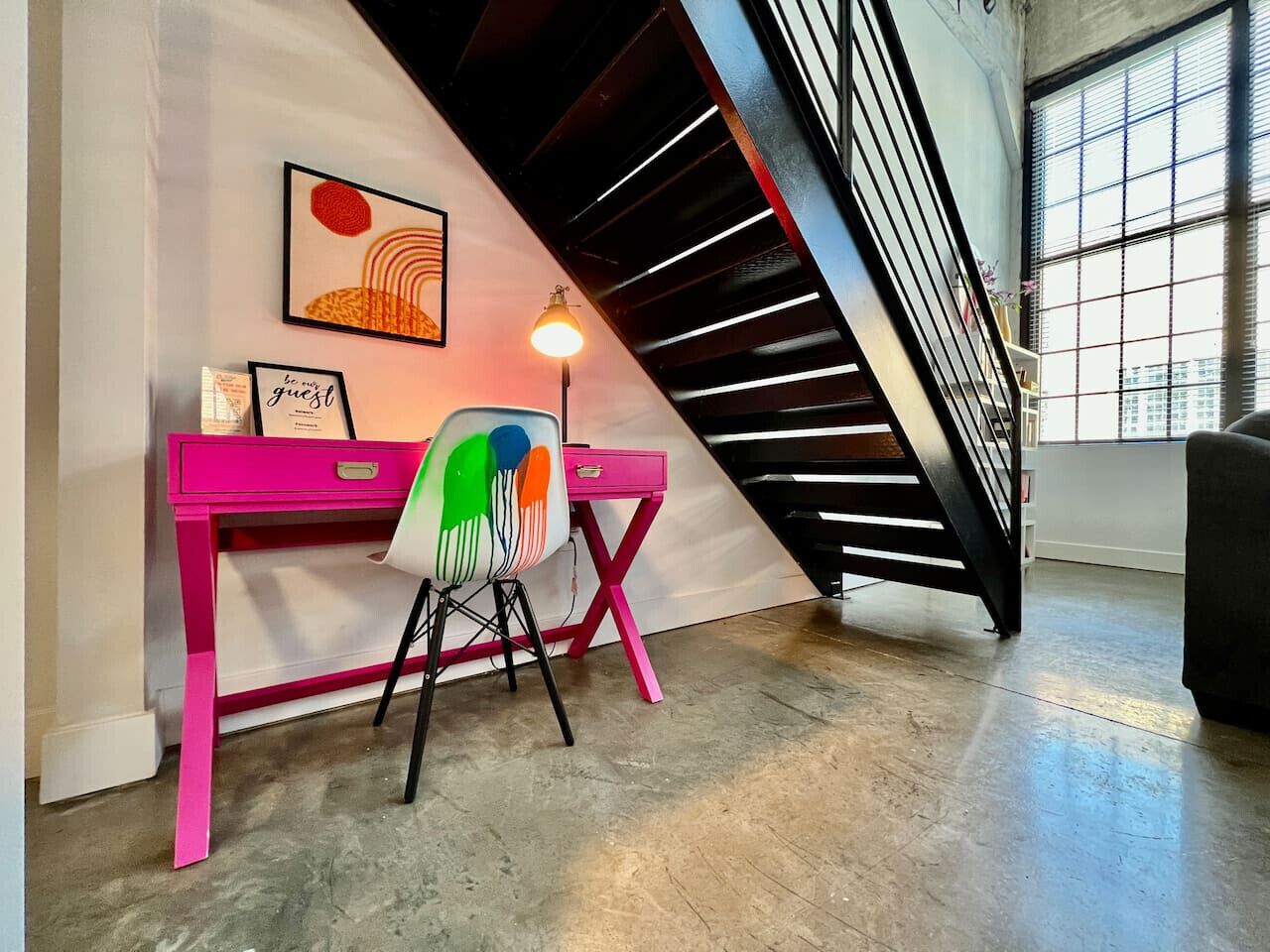 A room with a pink desk, a colorful chair, a lamp, and a staircase.