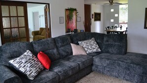 Ein Wohnzimmer mit einem grauen Ecksofa, einem roten Kissen und einem gemusterten Kissen. Im Hintergrund steht ein Esstisch mit Stühlen.