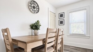Uma sala de jantar com uma mesa e cadeiras de madeira, um ventilador de teto, um relógio na parede e uma janela com persianas.