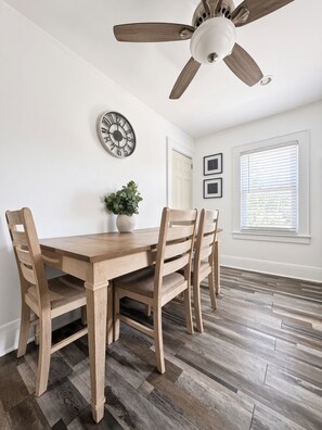 Une salle à manger avec une table et des chaises en bois, un ventilateur de plafond, une horloge murale et une fenêtre avec des stores.