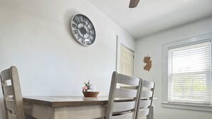 En matplats med ett träbord och stolar, en takfläkt, en väggklocka och ett fönster med persienner.