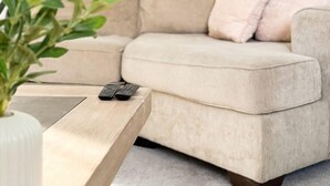 En beige soffa med en fjärrkontroll på ett träbord, en vit vas med gröna blad och ett konstverk på väggen.