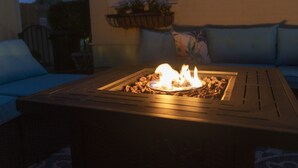 Mesa externa quadrada com lareira embutida, rodeada por um sofá azul e uma janela com um cesto de plantas.