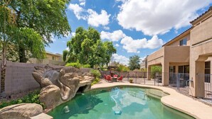 Una piscina con una formación rocosa, rodeada por una cerca y un patio con mobiliario exterior.