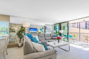 A modern living room with a sofa, coffee table, and a TV. There is a balcony with outdoor furniture.