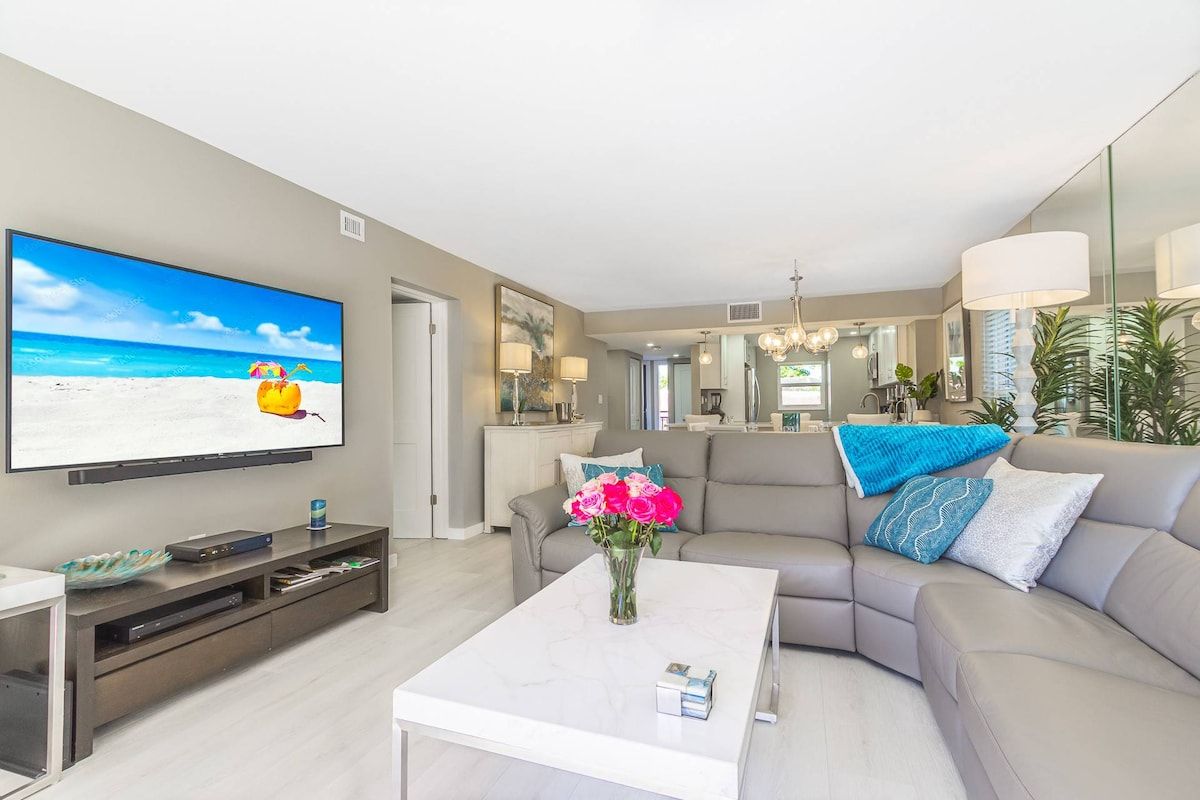 A modern living room with a flat-screen TV, a sofa, a coffee table, and a vase of flowers.