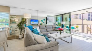 Un salon moderne avec un canapé, une table basse et un téléviseur. Il y a un balcon aménagé avec du mobilier d’extérieur.