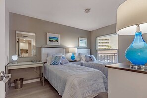 A hotel room with two beds, a vanity with a mirror, a blue lamp, and artwork on the walls.