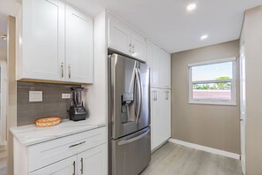A modern kitchen with white cabinets, a stainless steel refrigerator, a blender, and a window with a view.
