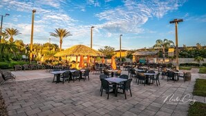 Un área de comedor al aire libre con mesas y sillas, rodeada de palmeras y una estructura con techo de paja.