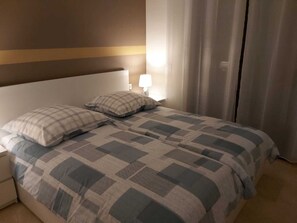A neatly made bed with a checkered bedspread, a bedside lamp, and a curtain on the window. - Top Ferienwohnung mit Pool in Spanien, Andalusien an der Costa del Sol (San Luis de Sabinillas)